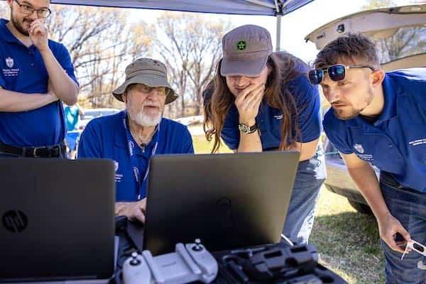 A male professor works on a laptop while three male students look on. The group is outside working under a tent and different equipment controllers can be seen in the foreground.