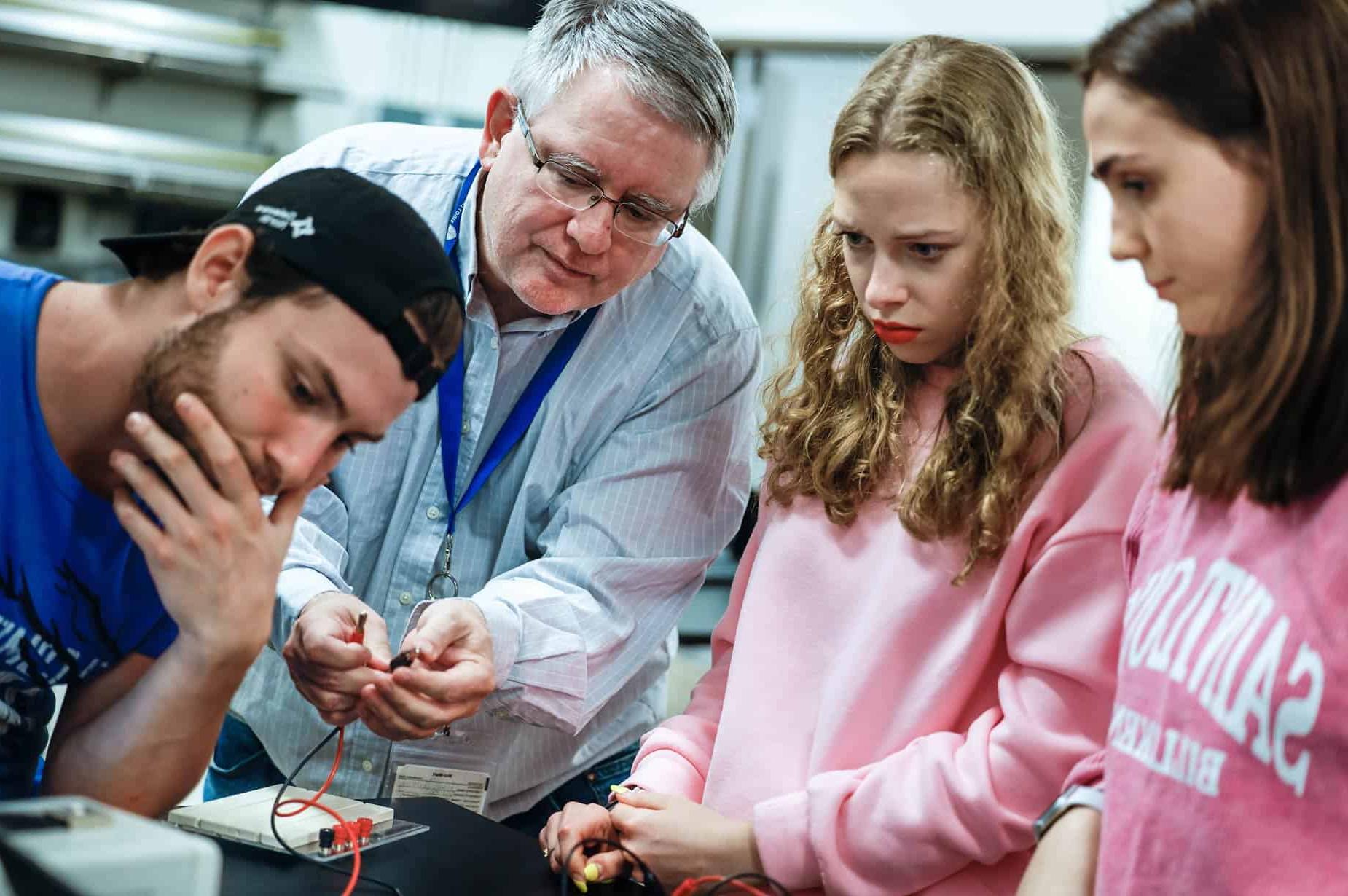 A male professor demonstrates how to use a piece of lab equipment to a group of three students. He holds a black and red wire in his hands.